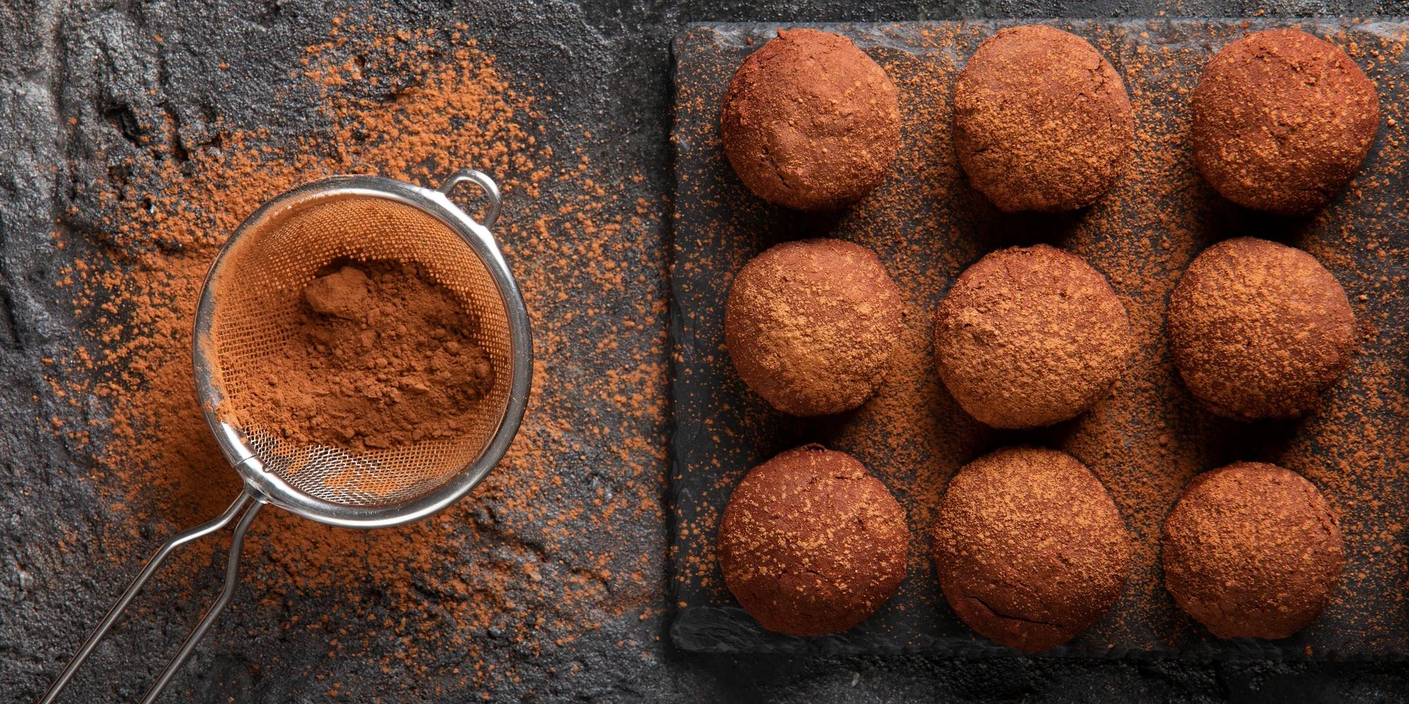Cocoa powder in a sieve beside a slate board of cocoa-dusted chocolate truffles
